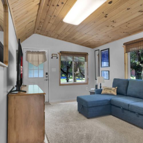 Living room with vaulted wooden ceiling, blue sectional sofa, and a cozy forest view through large windows.
