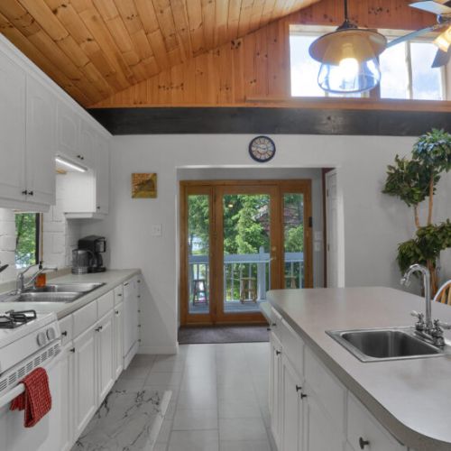 Bright, modern kitchen with white cabinetry, wood ceiling accents, and a view of the outdoor deck and forest.