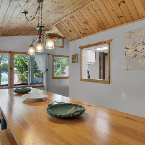 Bright dining area with natural wood finishes, pendant lighting, and a long table overlooking the greenery outside.
